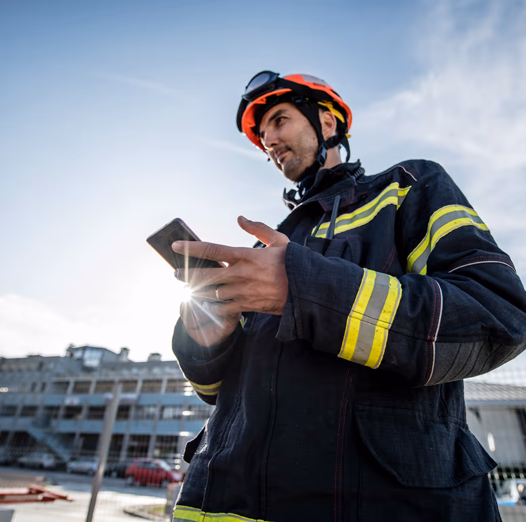 Firefighter in uniform and helmet using a smartphone outdoors, with the sun flaring behind him.