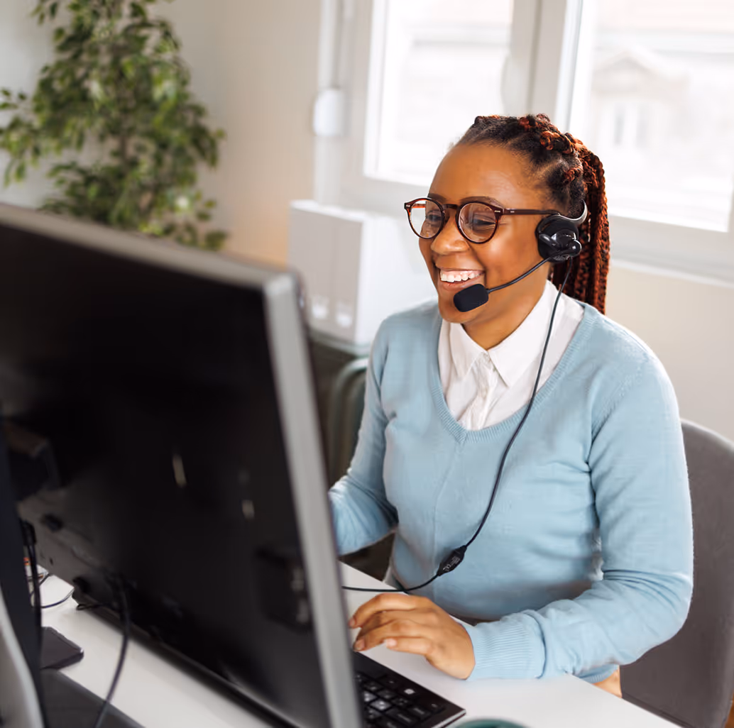 Smiling woman with glasses and headset working at a computer in a bright office.