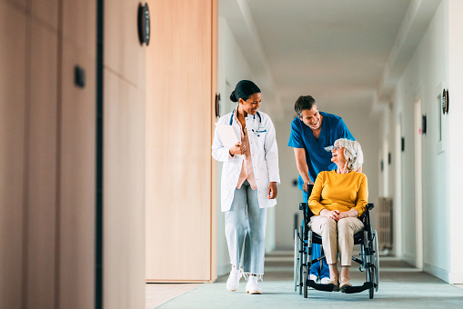 Doctor and nurse assisting an elderly patient in a wheelchair through a hospital corridor
